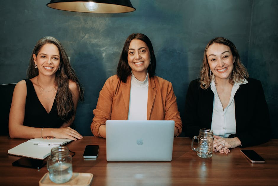 Three women in business attire collaborating at a table with laptops and notes, smiling confidently.